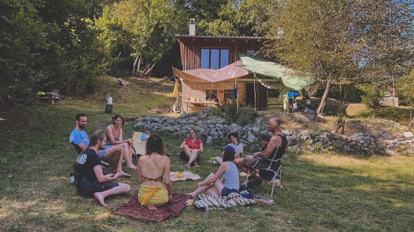 Gruppe sitzt im Kreis auf einer Wiese vor einer Holzhütte